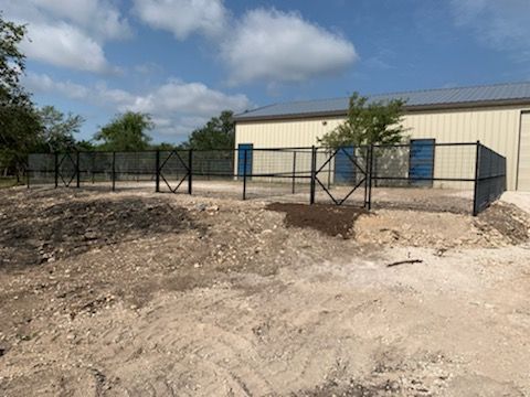Small beige building with blue doors behind a black metal fence on a sandy lot under a cloudy sky