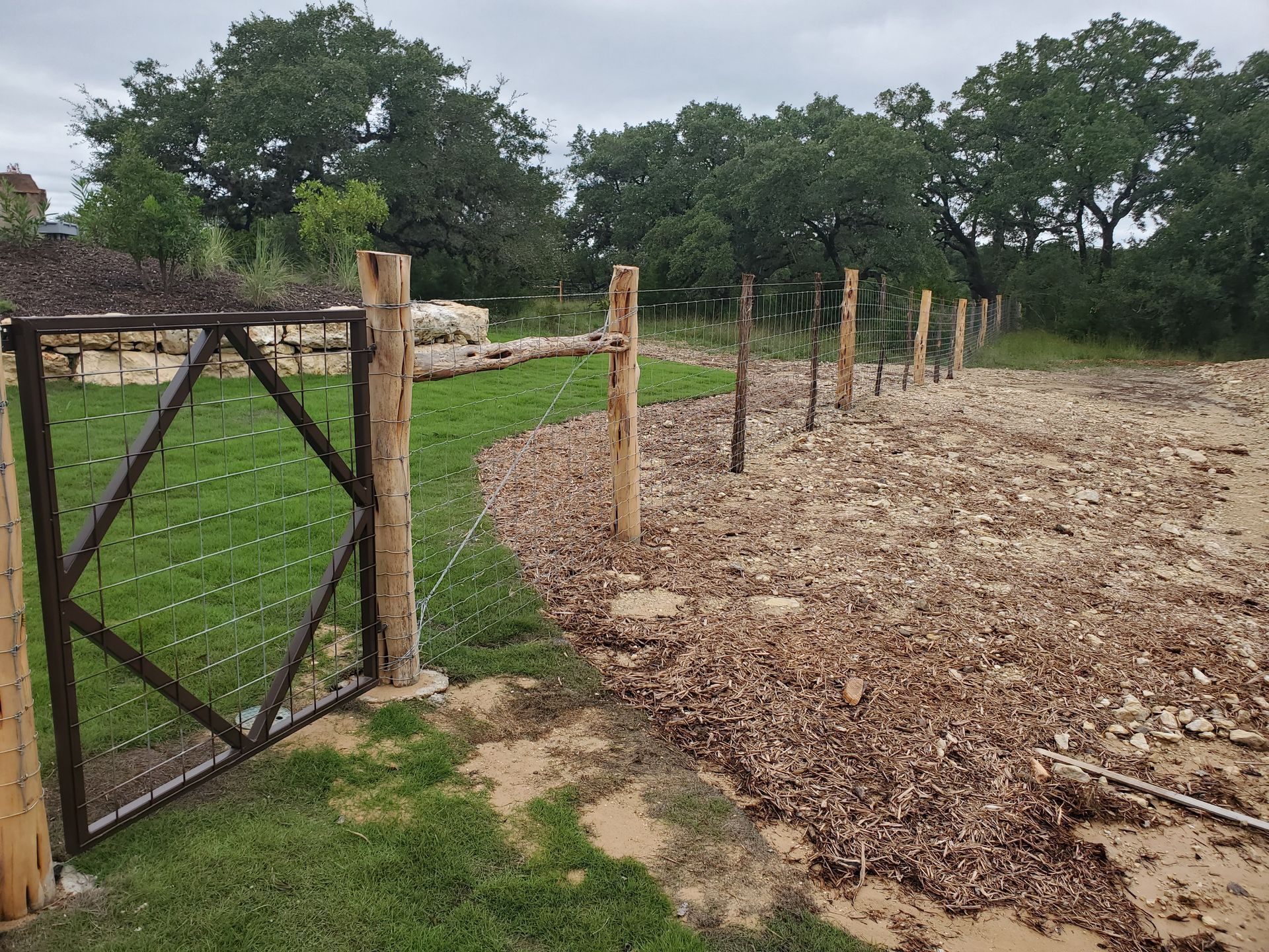 Fenced garden area with fresh dirt beds, wooden posts, and a rustic gate beside green grass and trees