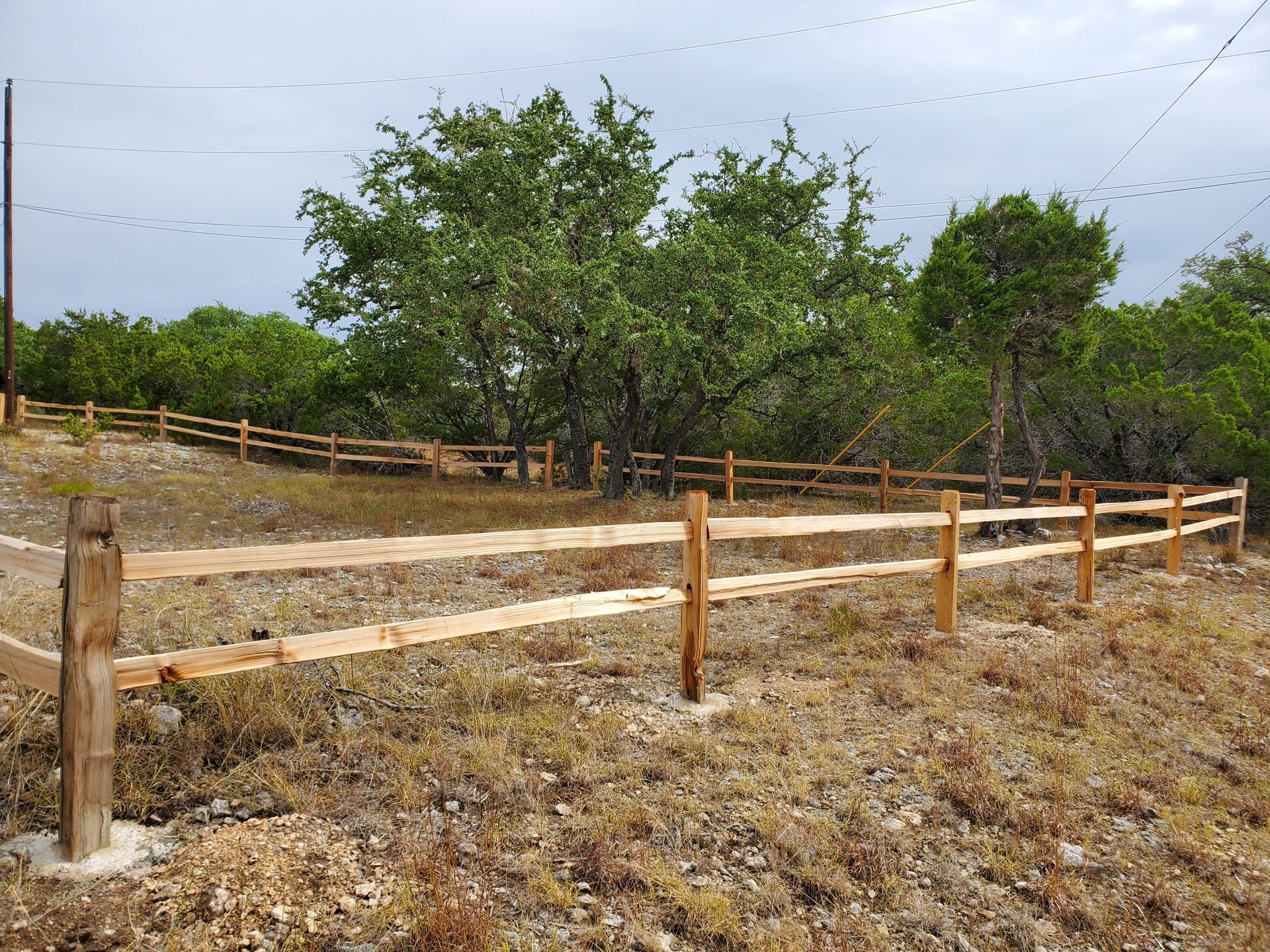 Wooden fence lining a dry dirt path beside green trees under a cloudy sky