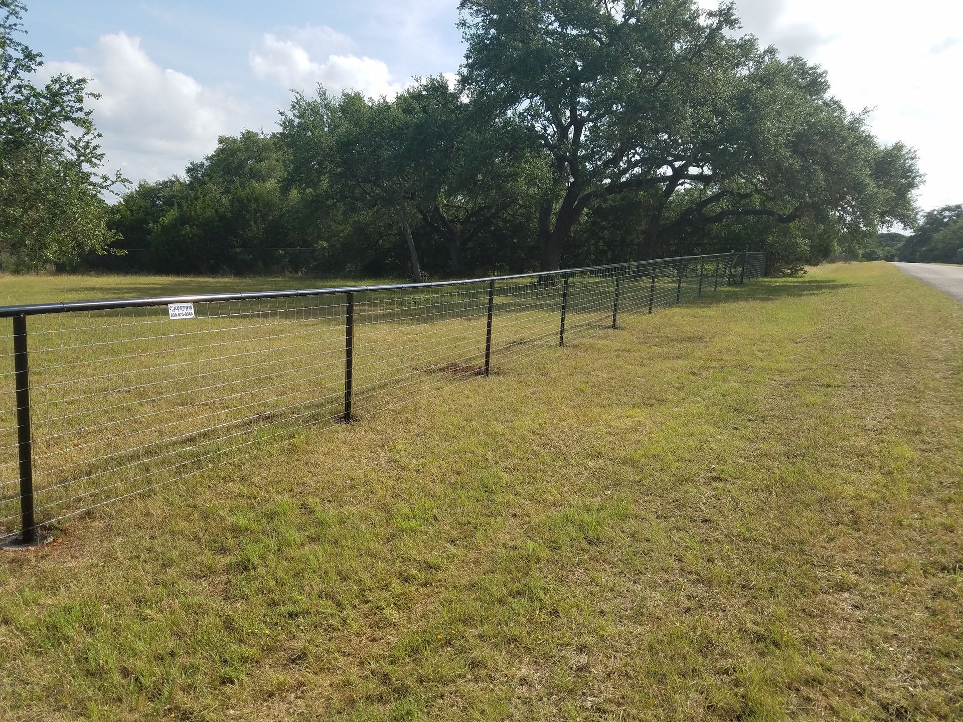 Black metal fence along a grassy roadside beside trees under a cloudy sky.