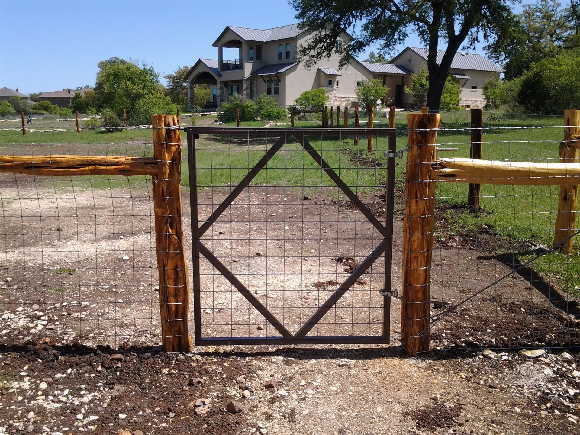 Rustic metal gate in front of a dirt path leading to a large house and trees.