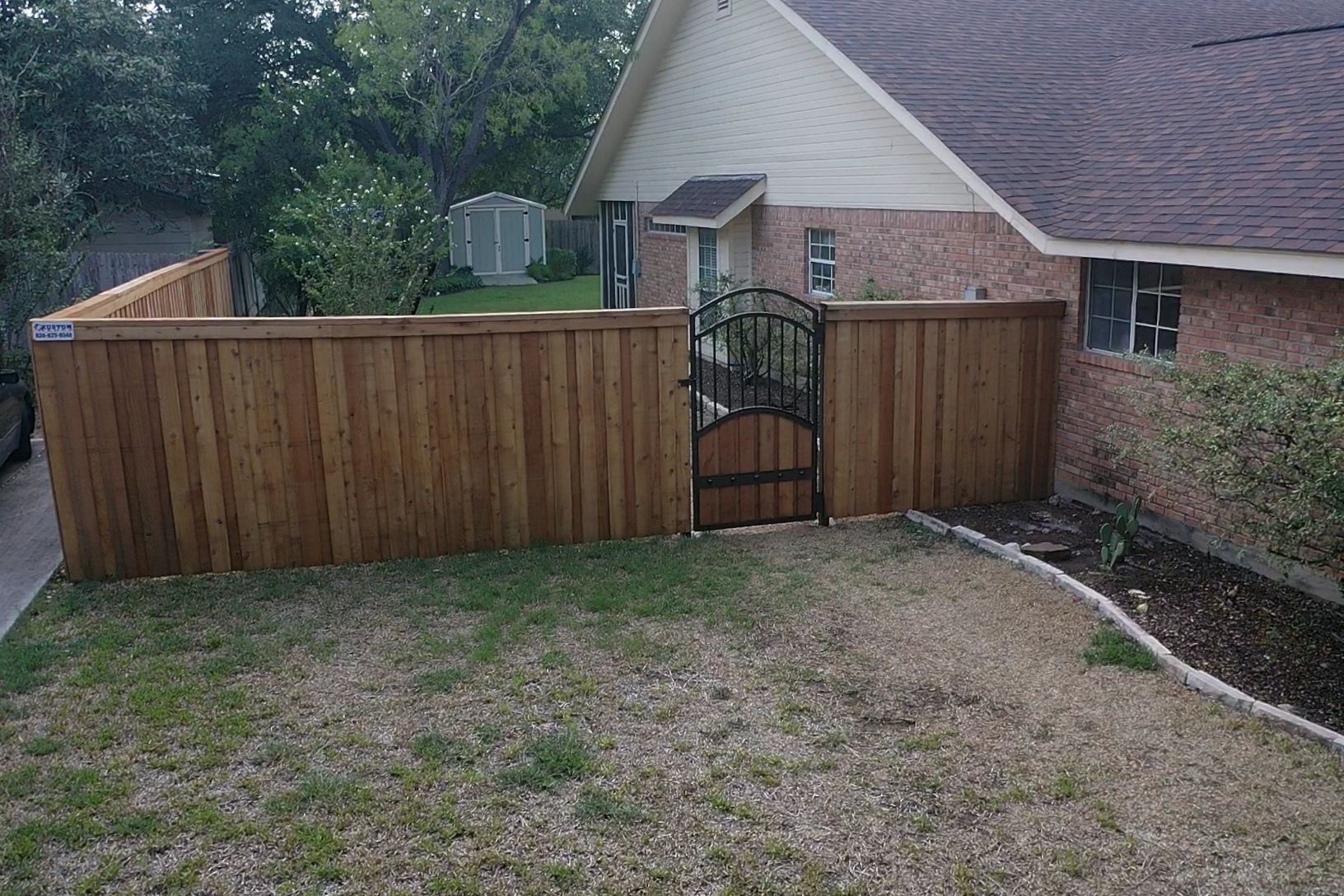 Wooden backyard fence with a black gate beside a brick house and sloped lawn.