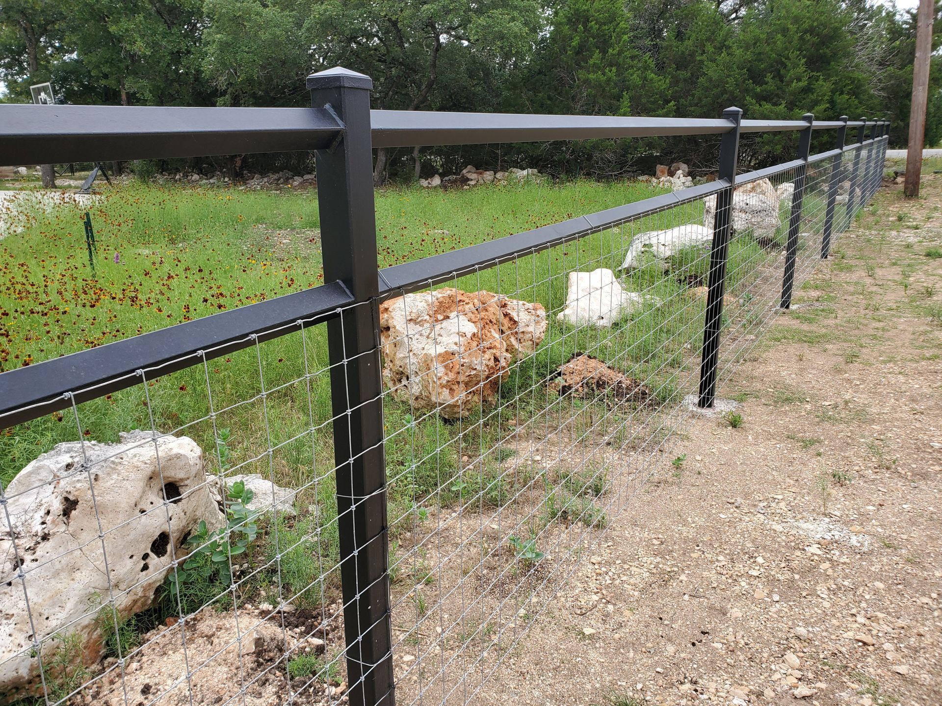 Black metal fence along a dirt path beside rocks and green grass in a wooded area.