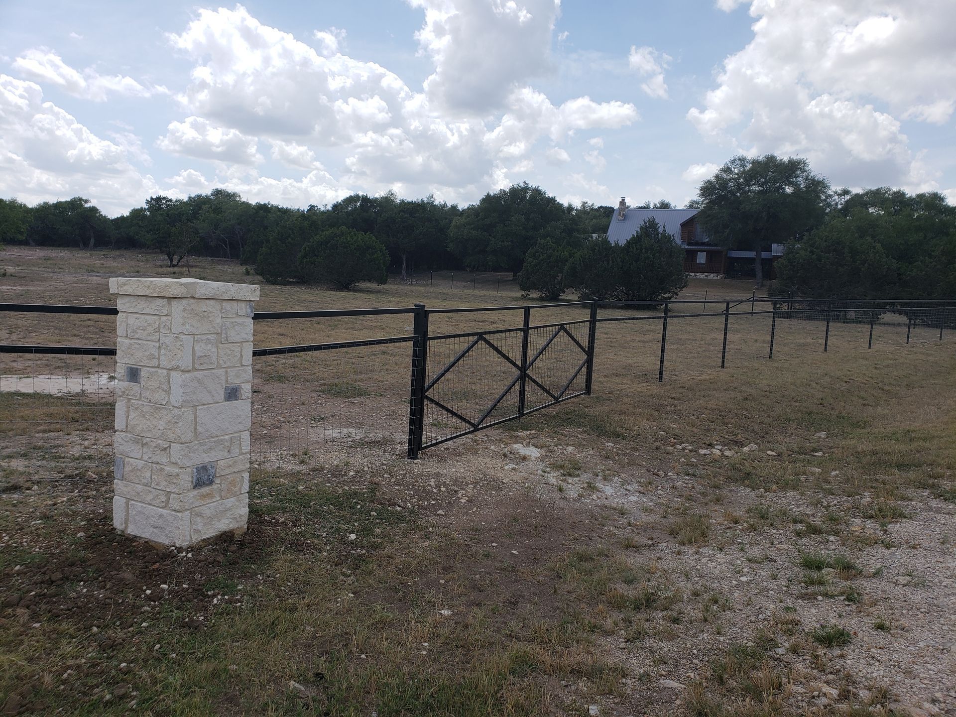 Stone fence post and black gate in a dry rural field under a cloudy sky, with trees and a house in the distance