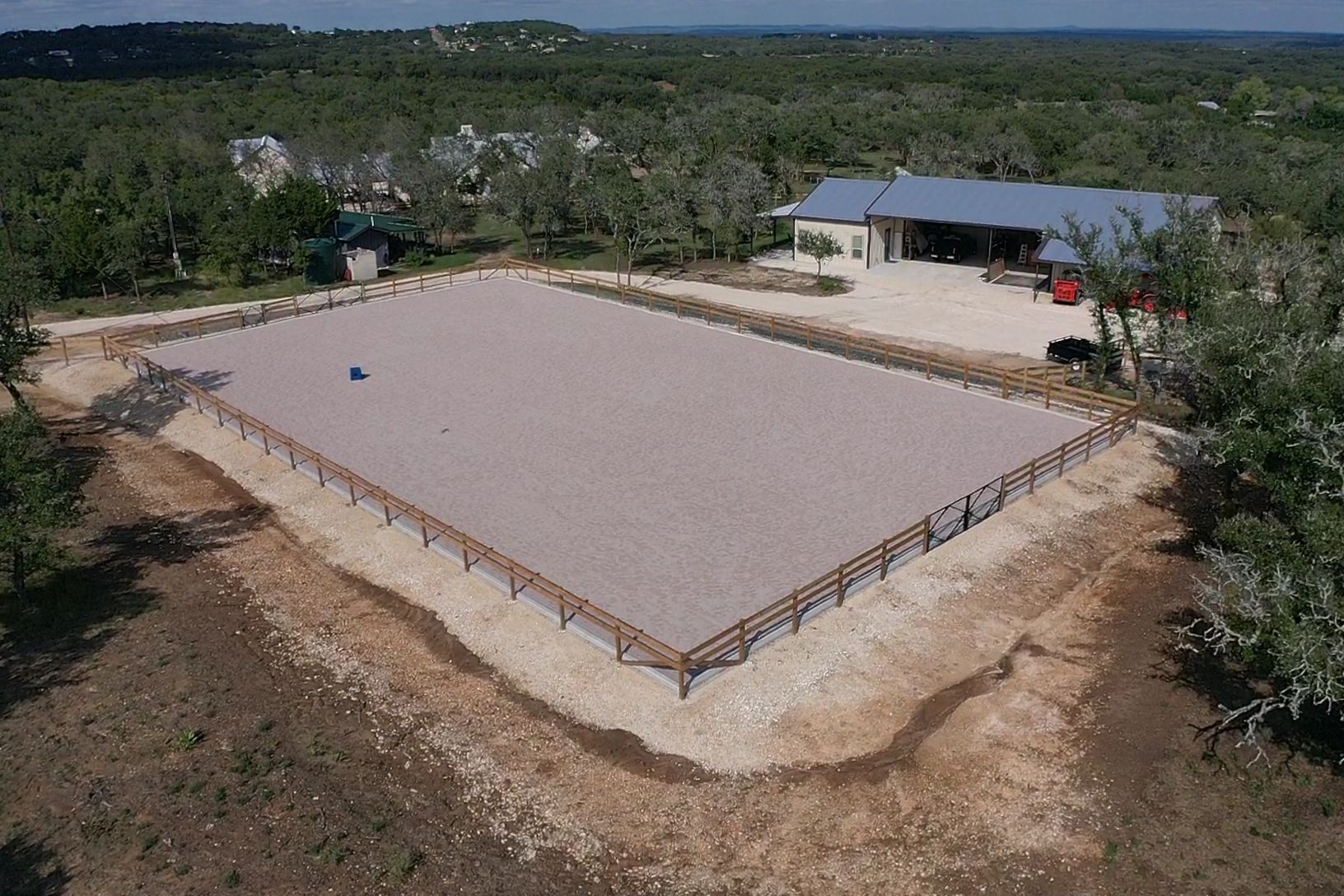 Large rectangular sand arena with low wooden rails, surrounded by trees and a small building in the background