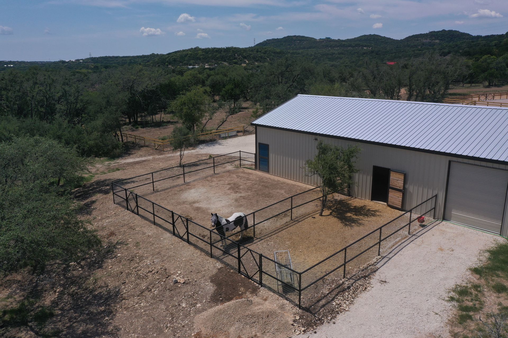 Rural barn with fenced pen and goats beside a gravel yard, surrounded by dry hills and trees