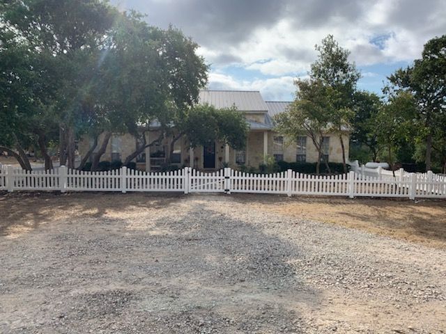 White house behind a white picket fence, with trees and a cloudy sky in front of a gravel yard