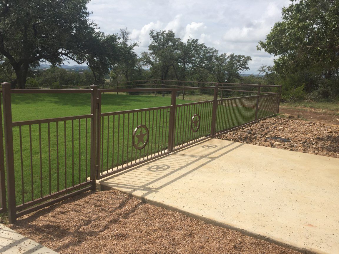 Brown metal gate beside a concrete driveway, opening to a green field under trees