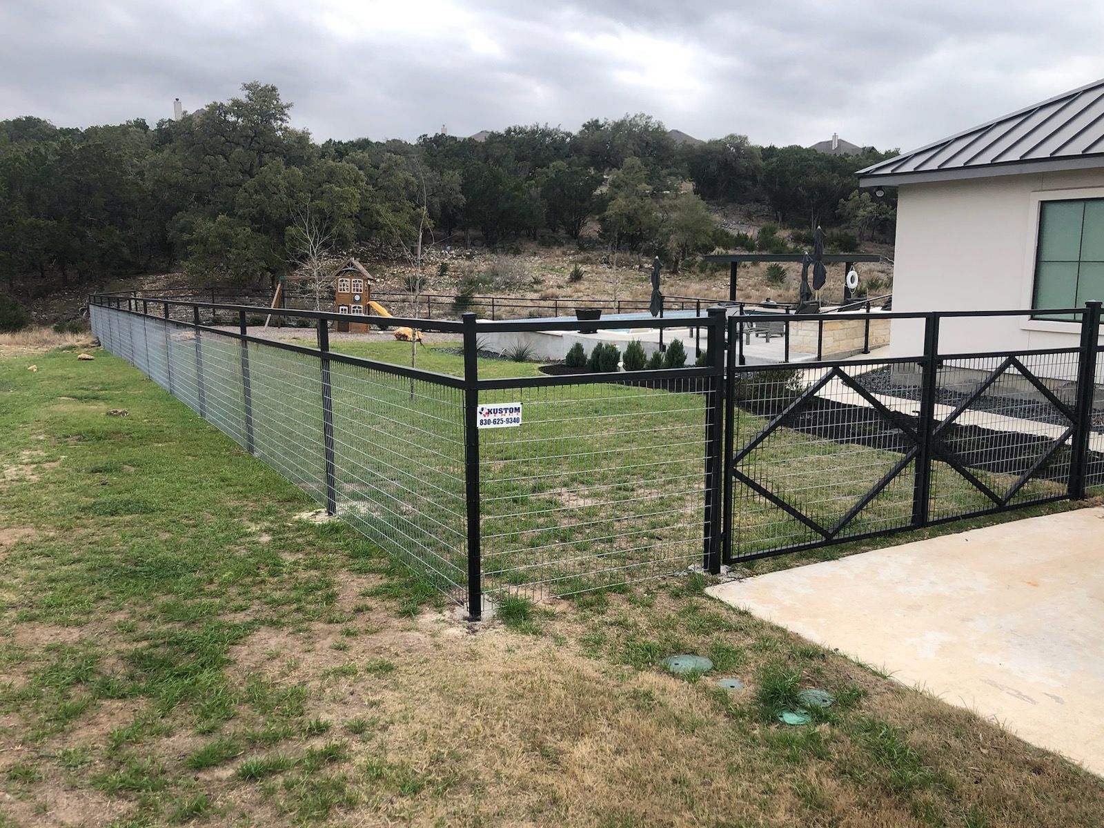 Chain-link fence and black gate beside a driveway, with a grassy yard and cloudy sky in the background.