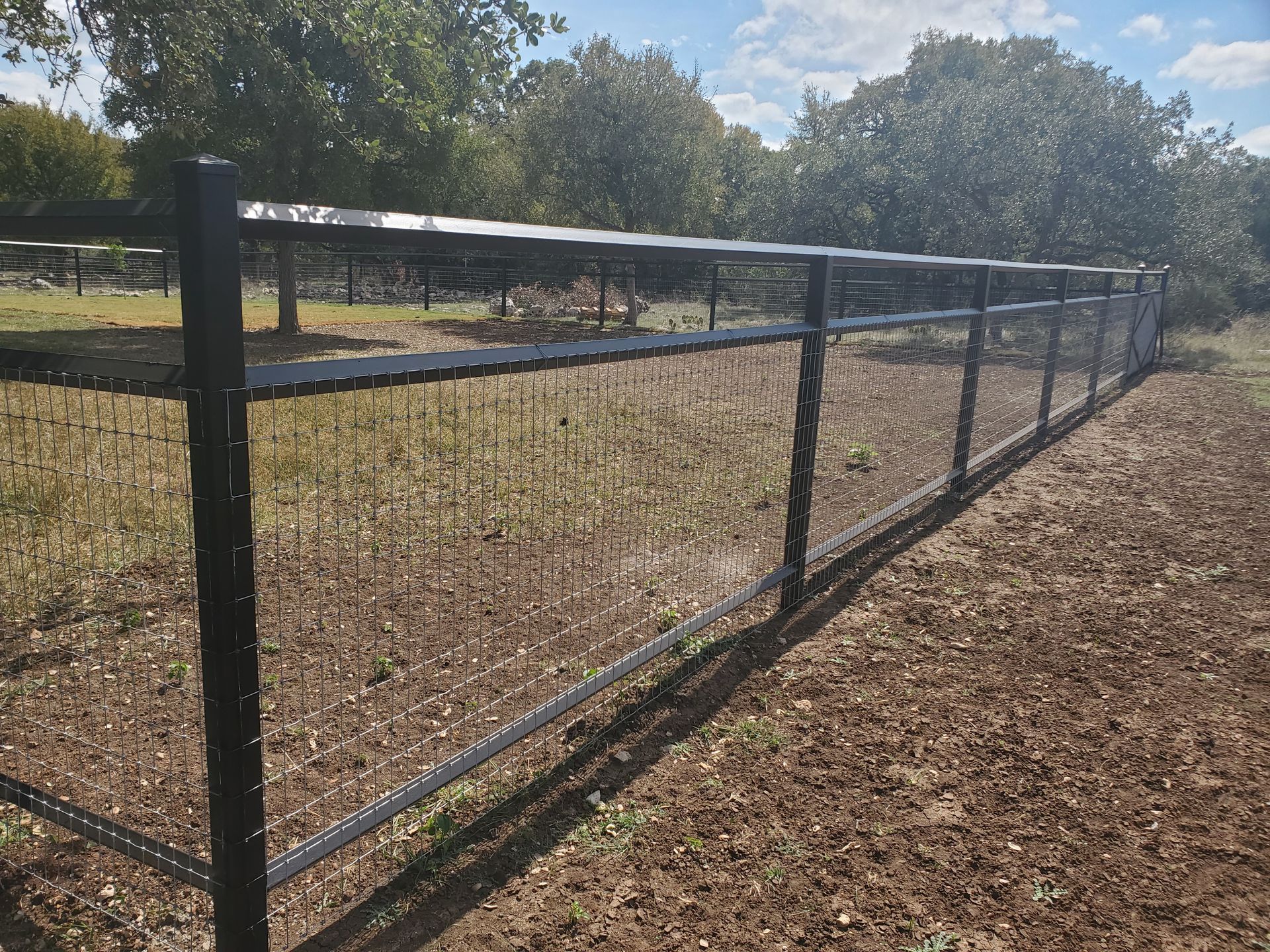 Black wire fence along a grassy field under trees on a sunny day