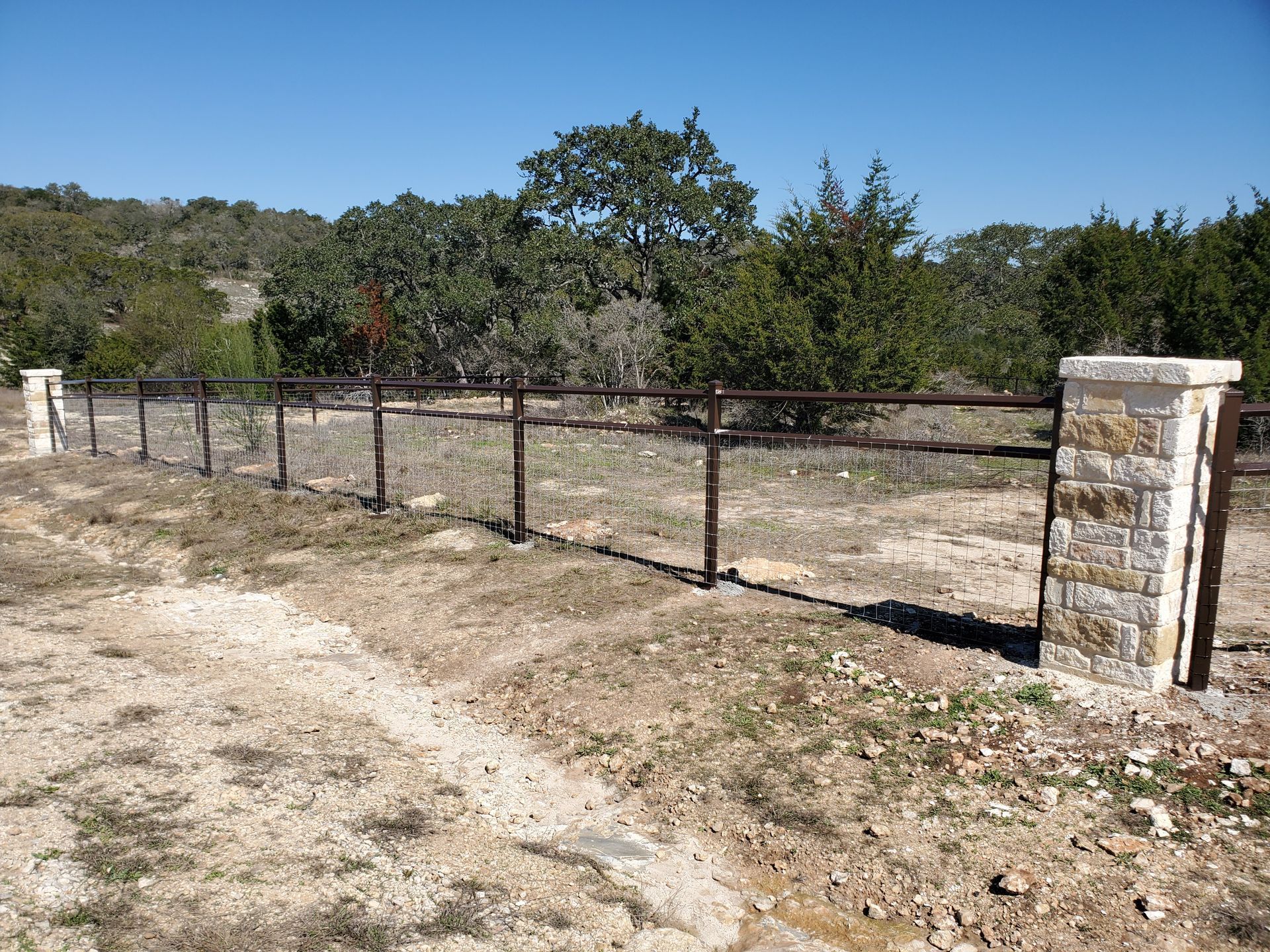 Rustic metal fence between stone pillars beside a dirt path in a dry, wooded area under a clear blue sky