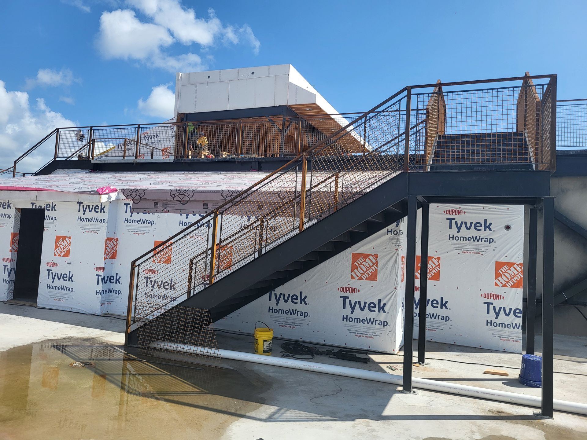 Exterior of a building under construction with a black metal staircase and Tyvek-wrapped walls under blue sky