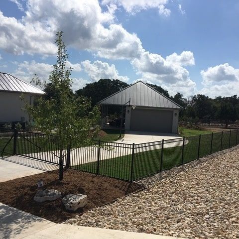 Front yard with young tree, black fence, gravel driveway, and gray garage under a cloudy sky