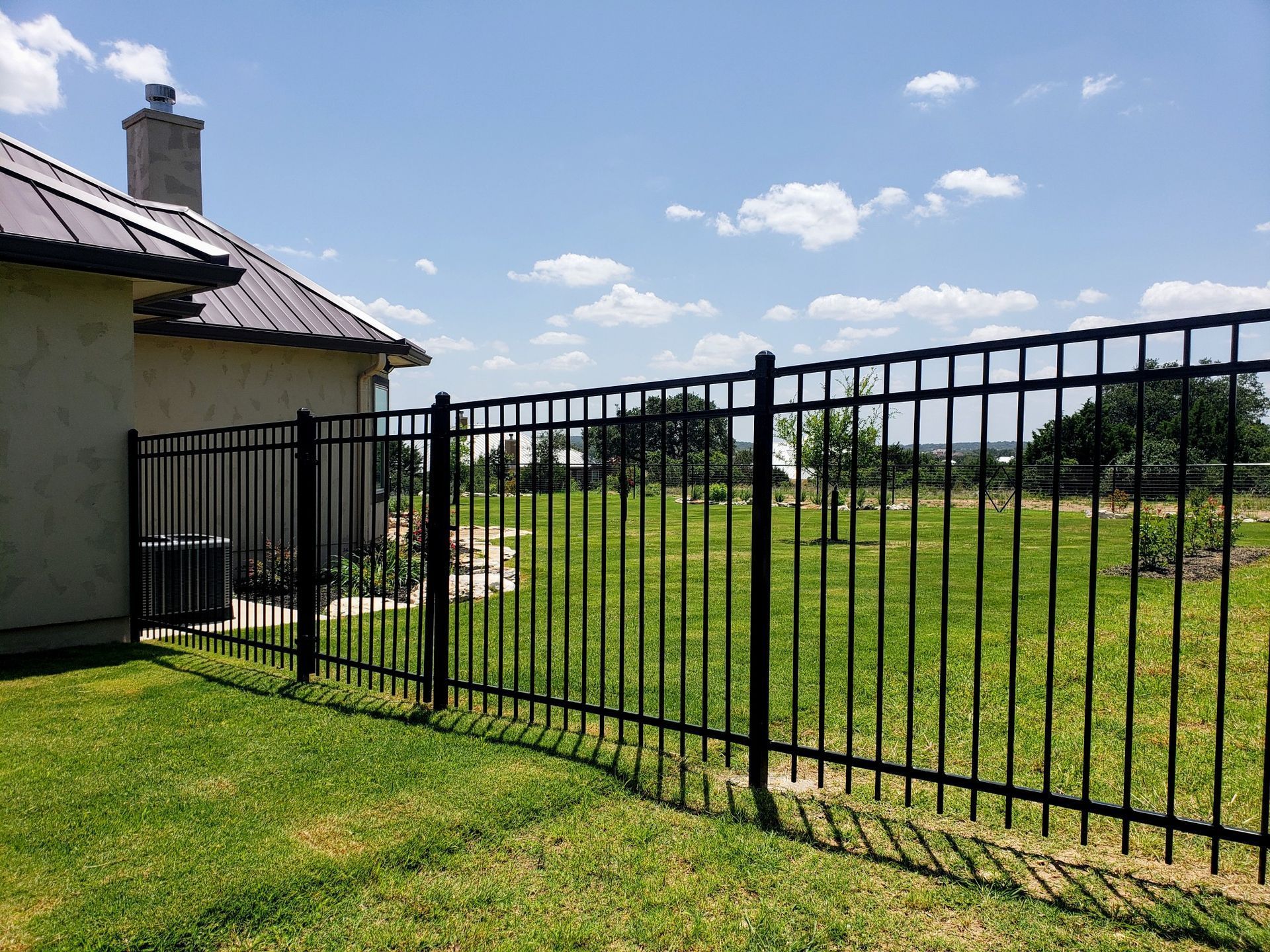 Black metal fence beside a house, bordering a grassy yard under a blue sky