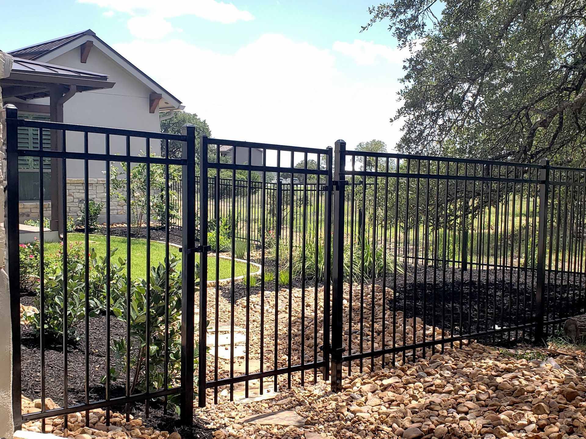 Black metal fence with gate beside a house and garden beds under a partly cloudy sky