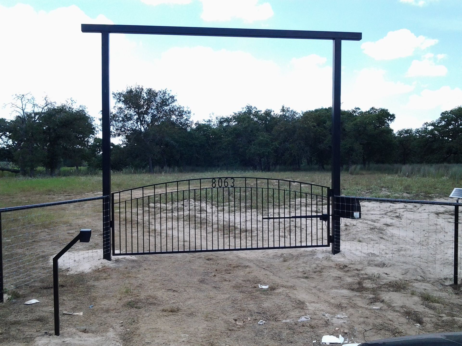 Metal gate on a dirt path in a grassy field, framed by two black posts under a cloudy sky.