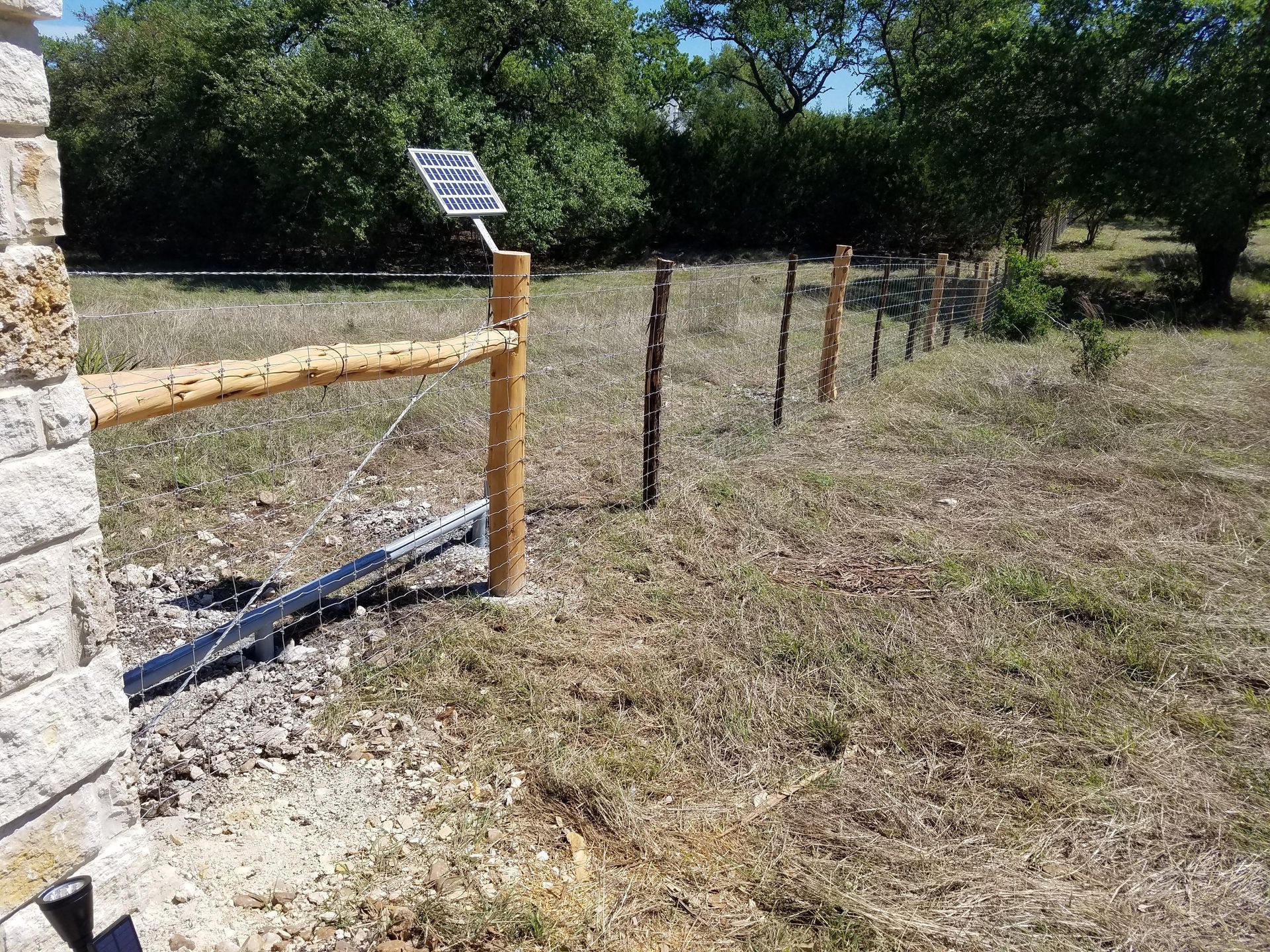 Rustic fence line beside a stone wall in a sunny grassy field with trees in the background