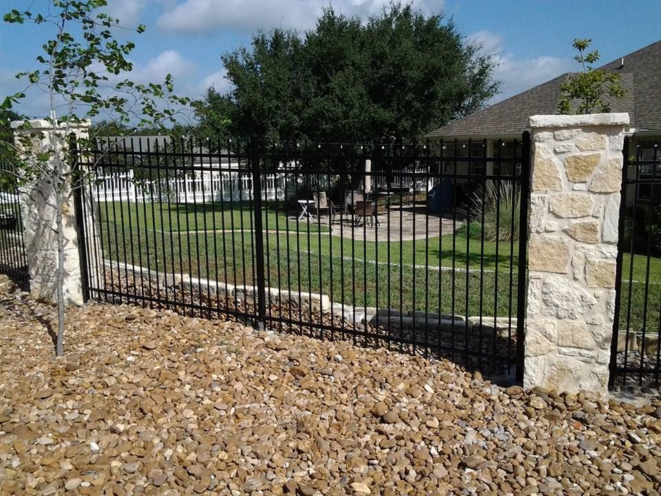 Black metal gate between stone pillars in a front yard with lawn and trees behind it.