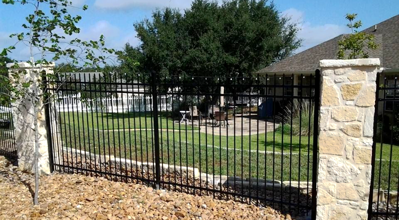 Black metal gate between stone pillars in a landscaped yard with a tree and house in the background