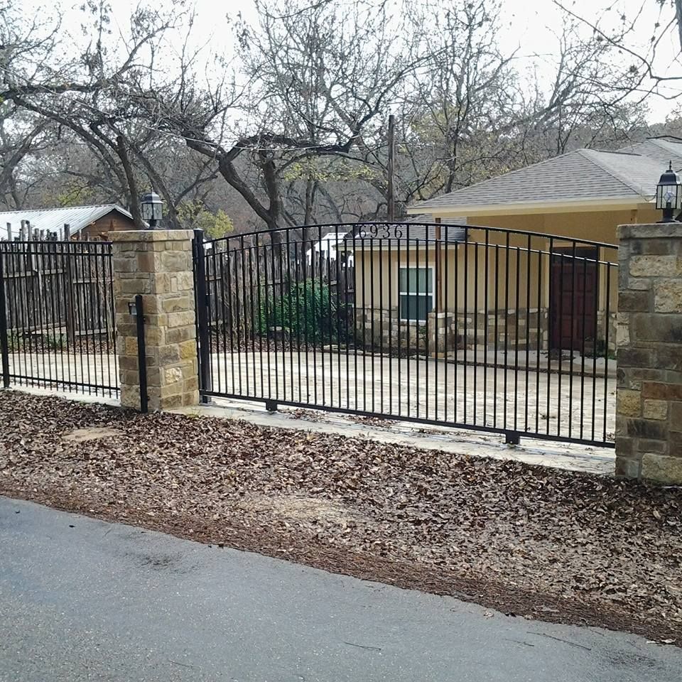 Black metal gate and stone fence in front of a tan house on a quiet street.