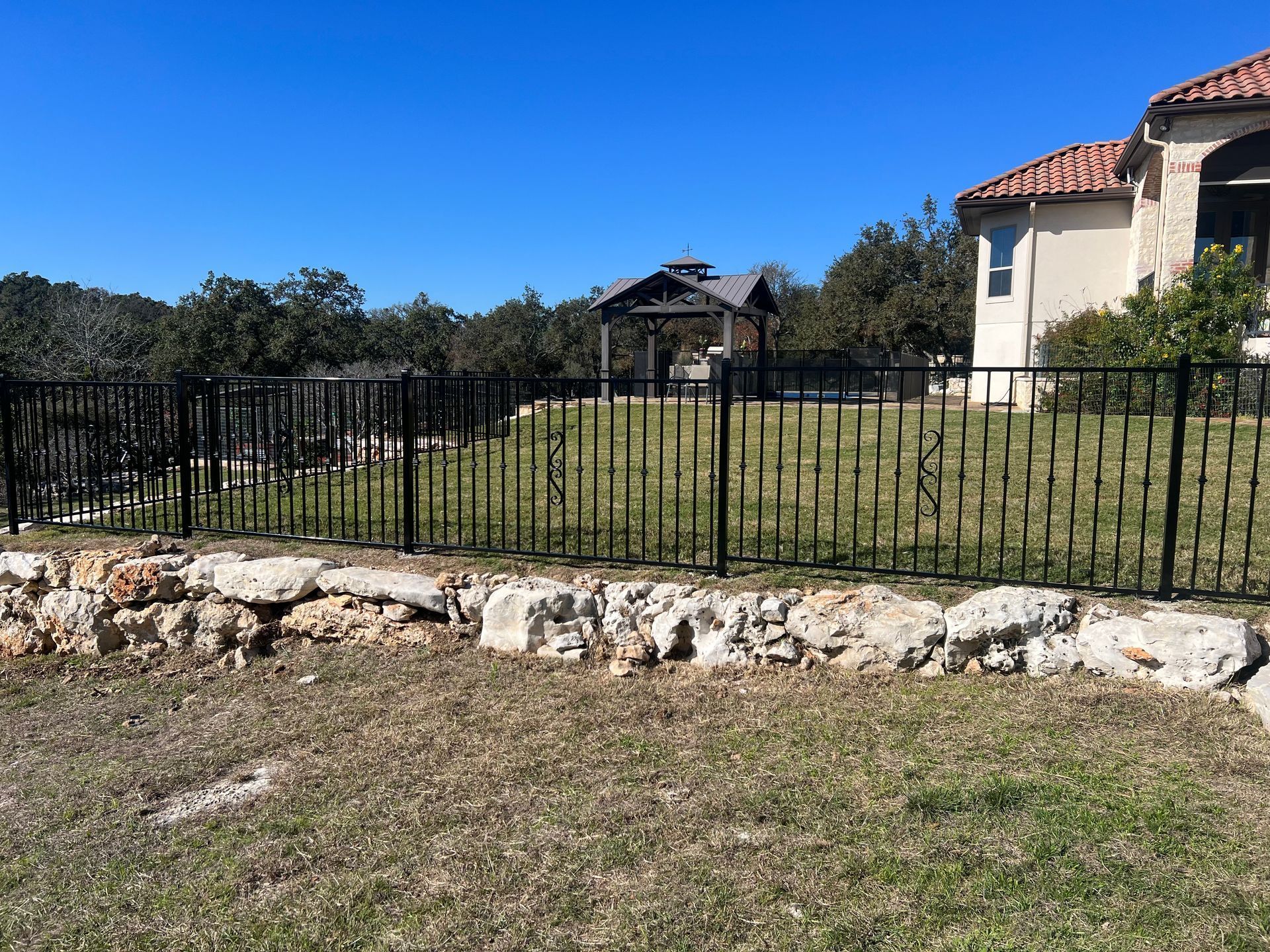 Black metal fence along a grassy yard with stone border, houses in the background, and blue sky.