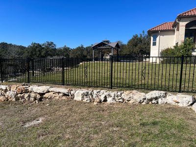 Black metal fence along a grassy yard with stone border, houses in the background, and blue sky.