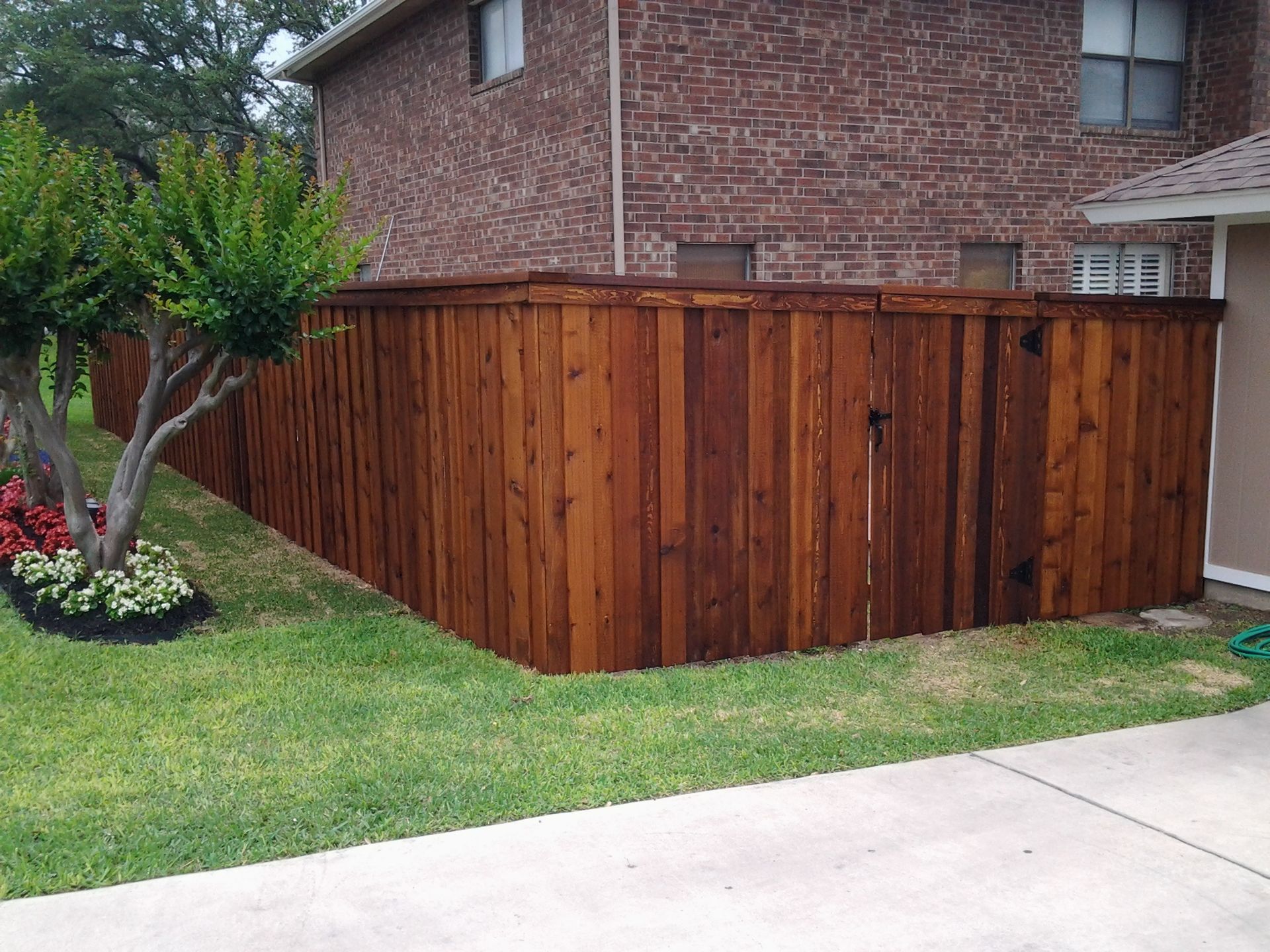 Wooden privacy fence beside a brick house, bordering a green lawn and sidewalk.