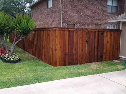 Wooden privacy fence beside a brick house, bordering a green lawn and sidewalk.
