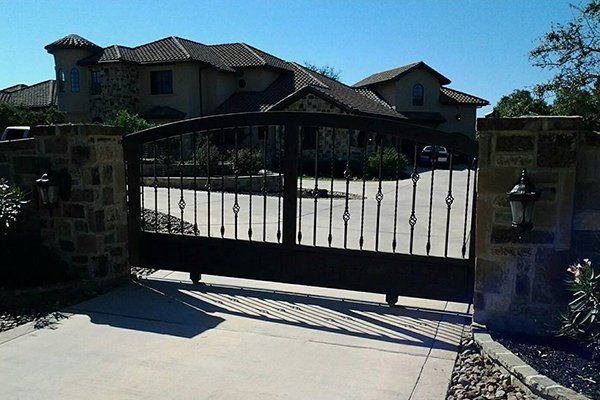 Black metal driveway gate in front of a large house, with stone pillars and bright sunlight