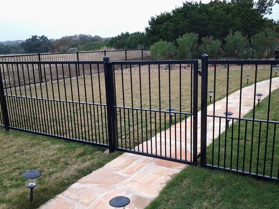 Black metal fence with a gate across a grassy yard and stone walkway, with trees and open field beyond