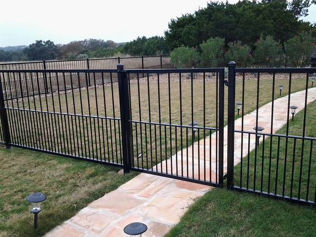 Black metal fence with a gate across a grassy yard and stone walkway, with trees and open field beyond