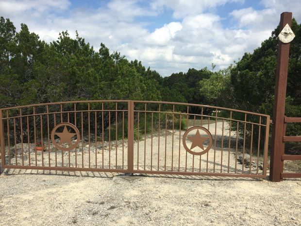 Brown metal gate with star cutouts blocking a gravel driveway, framed by trees and a cloudy sky.