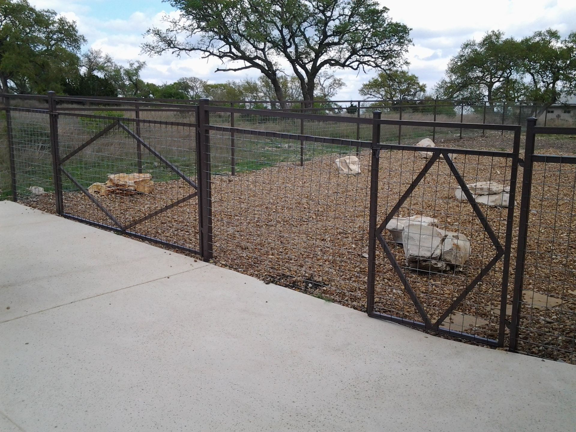 Black metal fence around gravel area with a small opening beside a concrete sidewalk and trees beyond