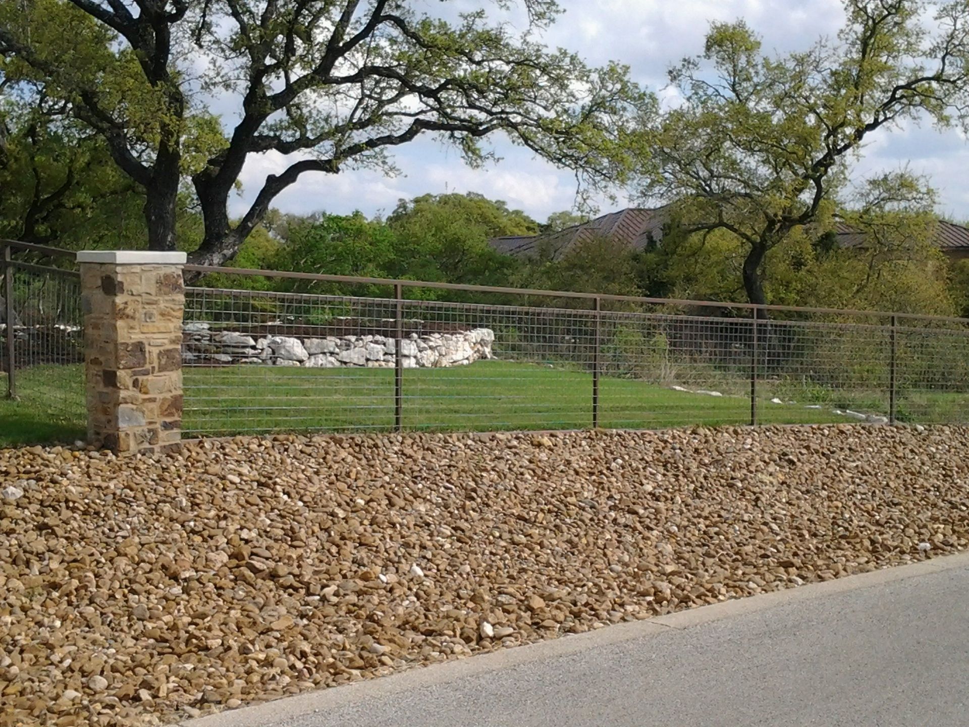Stone wall and metal fence beside a rocky roadside, with green grass and trees under a cloudy sky