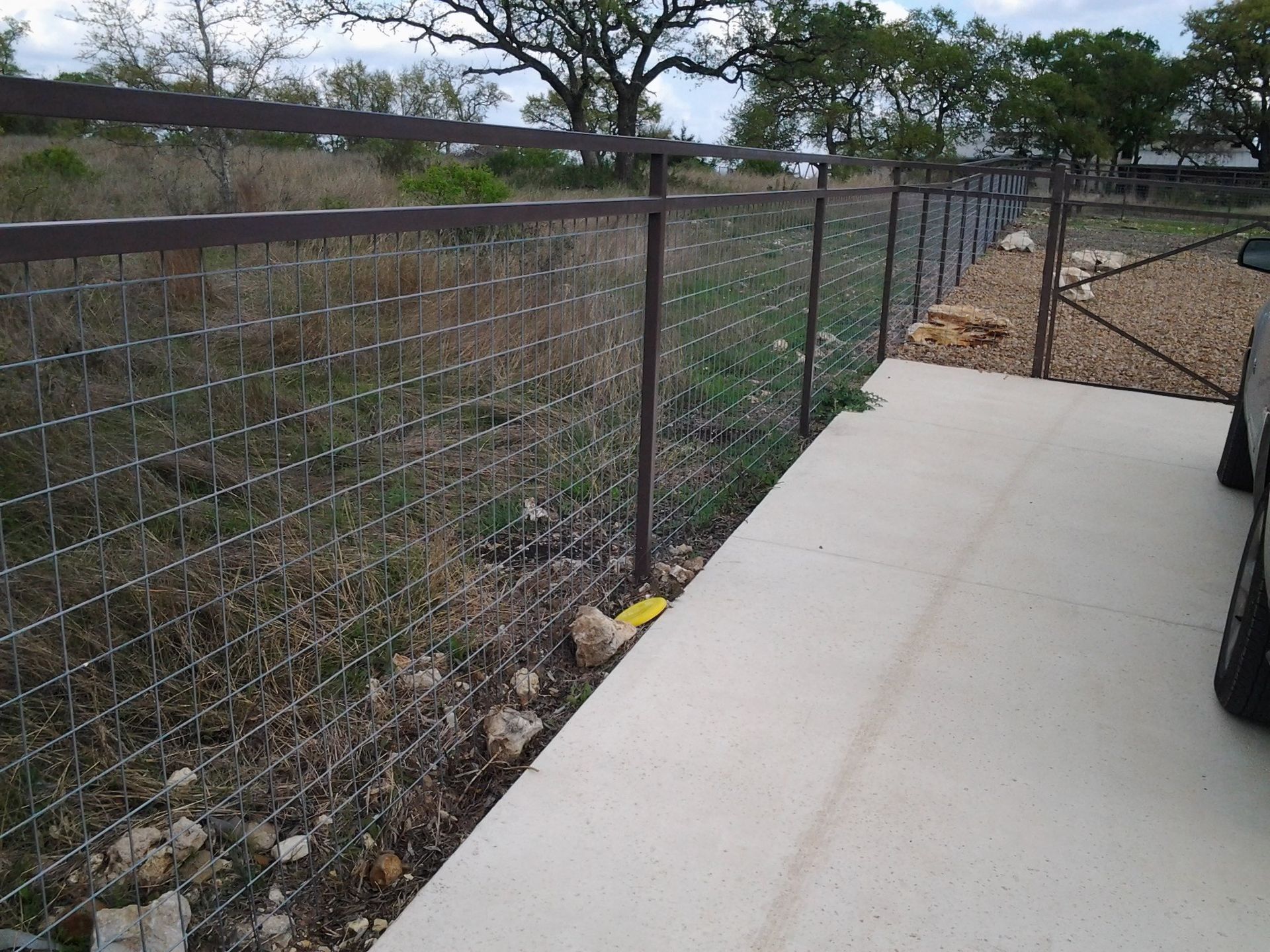 Concrete walkway beside a chain-link fence along a dirt embankment with trees in the background.