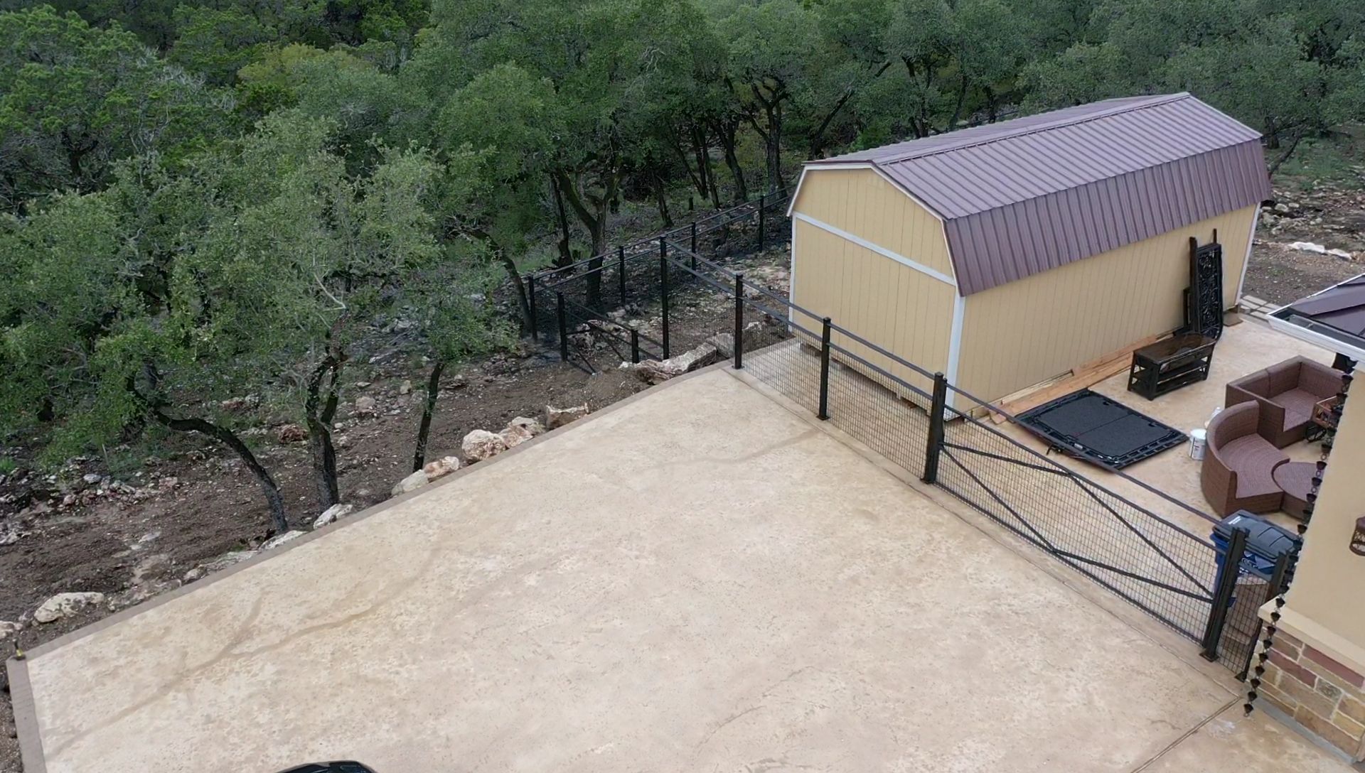 Patio beside a tan shed overlooking a wooded hillside, with a fence and stacked firewood.