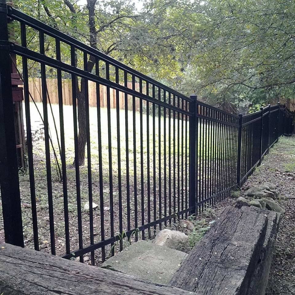Black metal fence beside a wooded slope with a concrete path and fallen logs on the ground