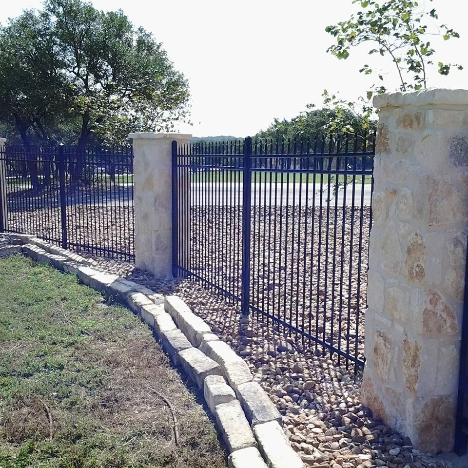 Blue metal fence with stone pillars beside a gravel road and grassy area under trees