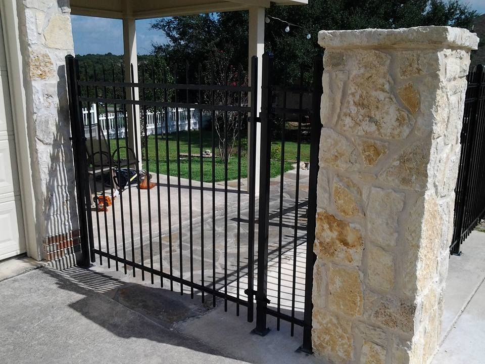 Black metal gate between stone pillars at a driveway entrance, with patio and lawn beyond.