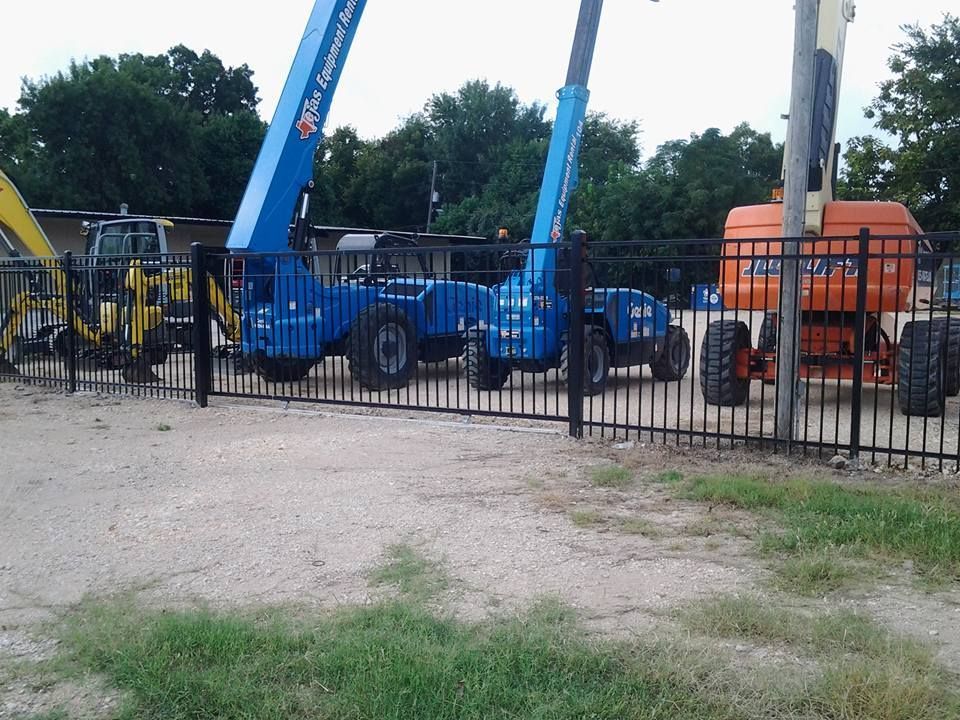 Blue and orange construction lifts behind a black fence on a gravel lot with trees in the background