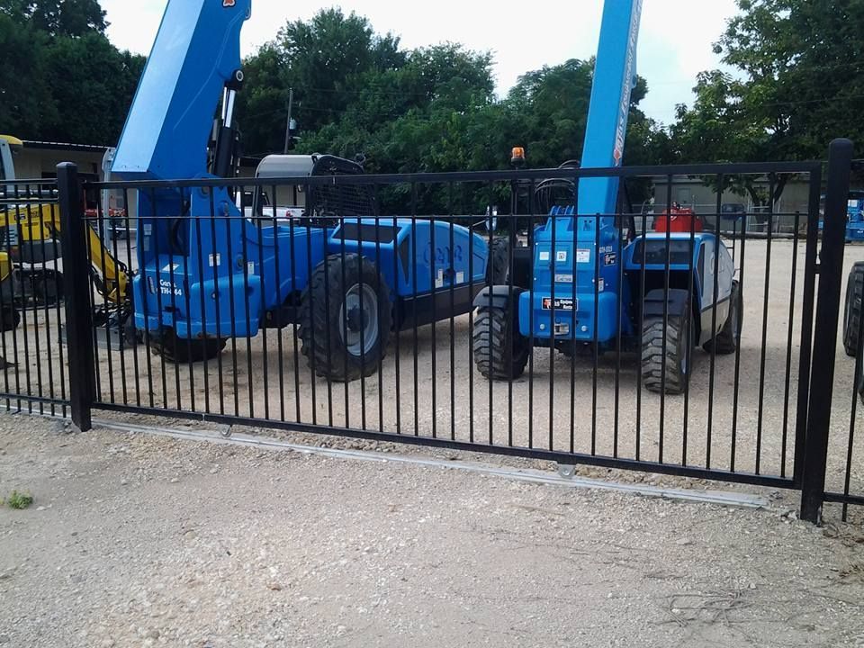 Blue construction lift and telescopic handler behind a black fence on a gravel lot.