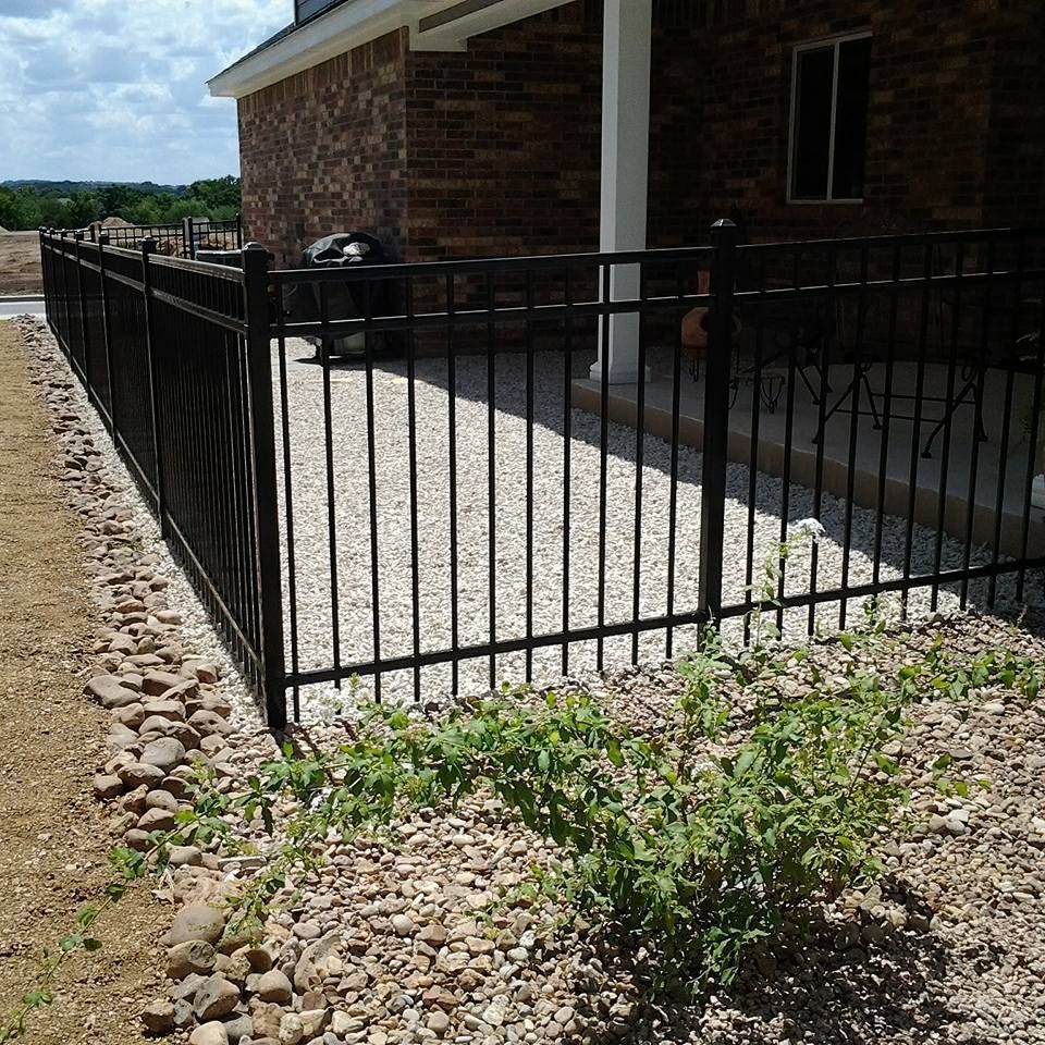 Black metal fence around a gravel patio beside a brick house and rock landscaping under a sunny sky