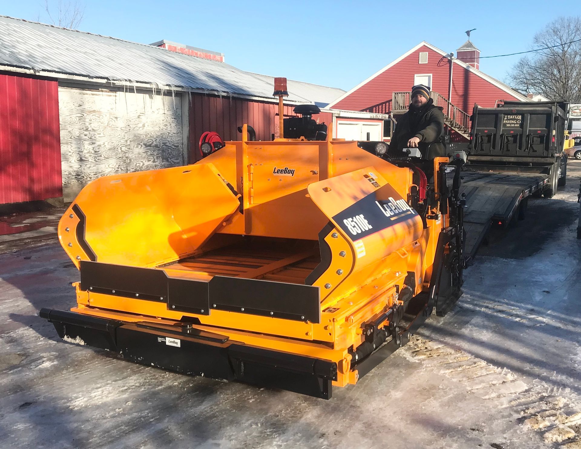 A man is driving an orange asphalt plow in front of a red barn.
