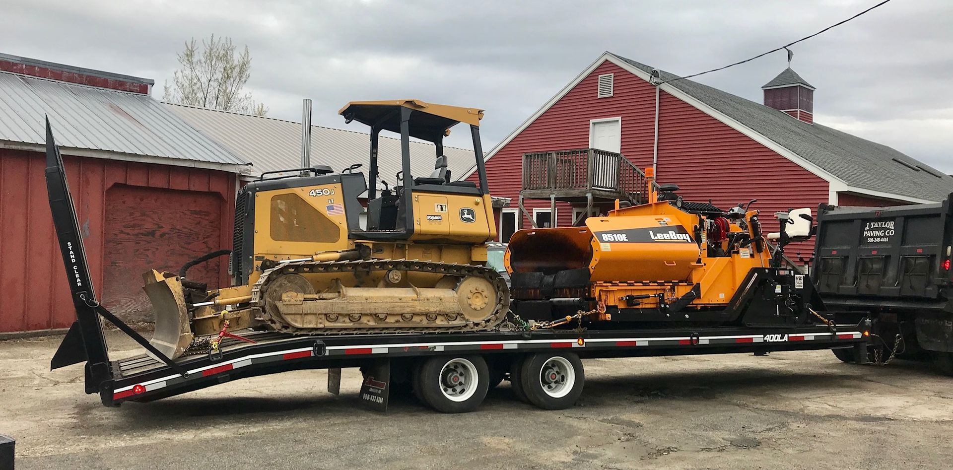 A bulldozer is sitting on top of a trailer in front of a red barn.