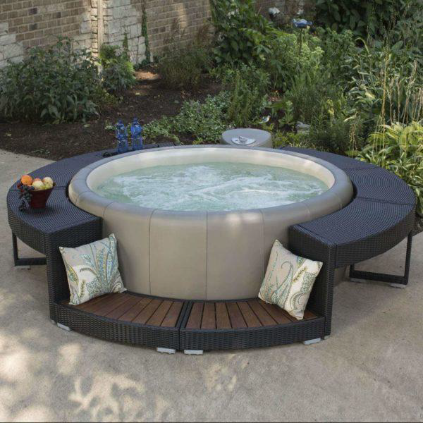 Circular hot tub with surrounding dark wicker benches, cushions, and small table. Lush greenery in background.