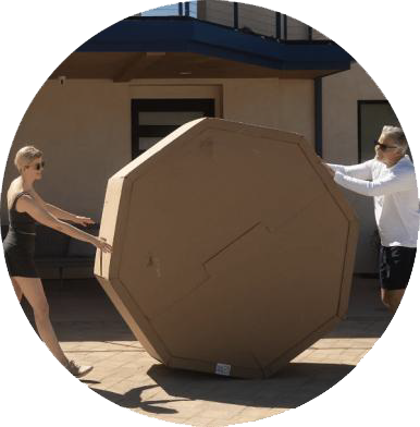 Two people rolling a large, octagonal cardboard box outdoors.