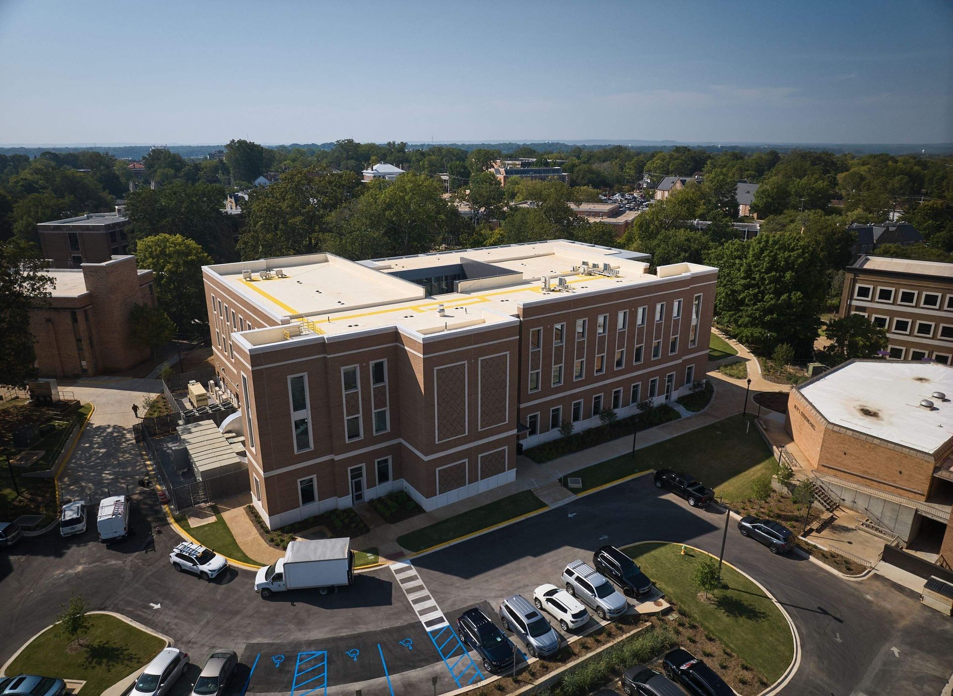 Aerial view of a brick building with parked cars and surrounding trees on a sunny day