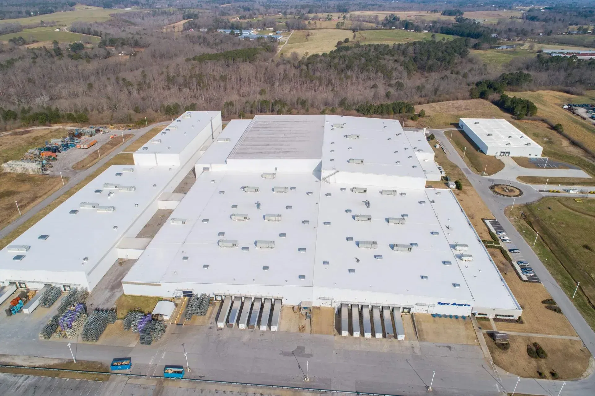 Aerial view of a large white industrial warehouse with loading docks and trucks in a rural area