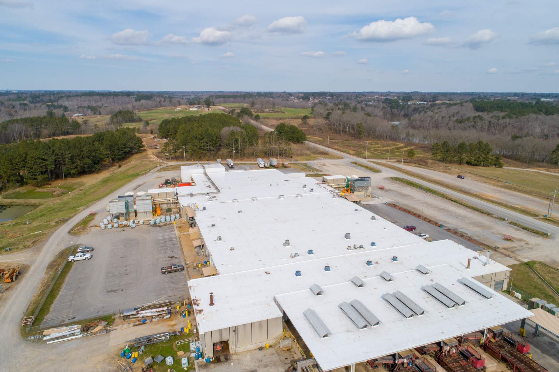 Aerial view of a large white industrial building under construction beside a parking lot and trees.