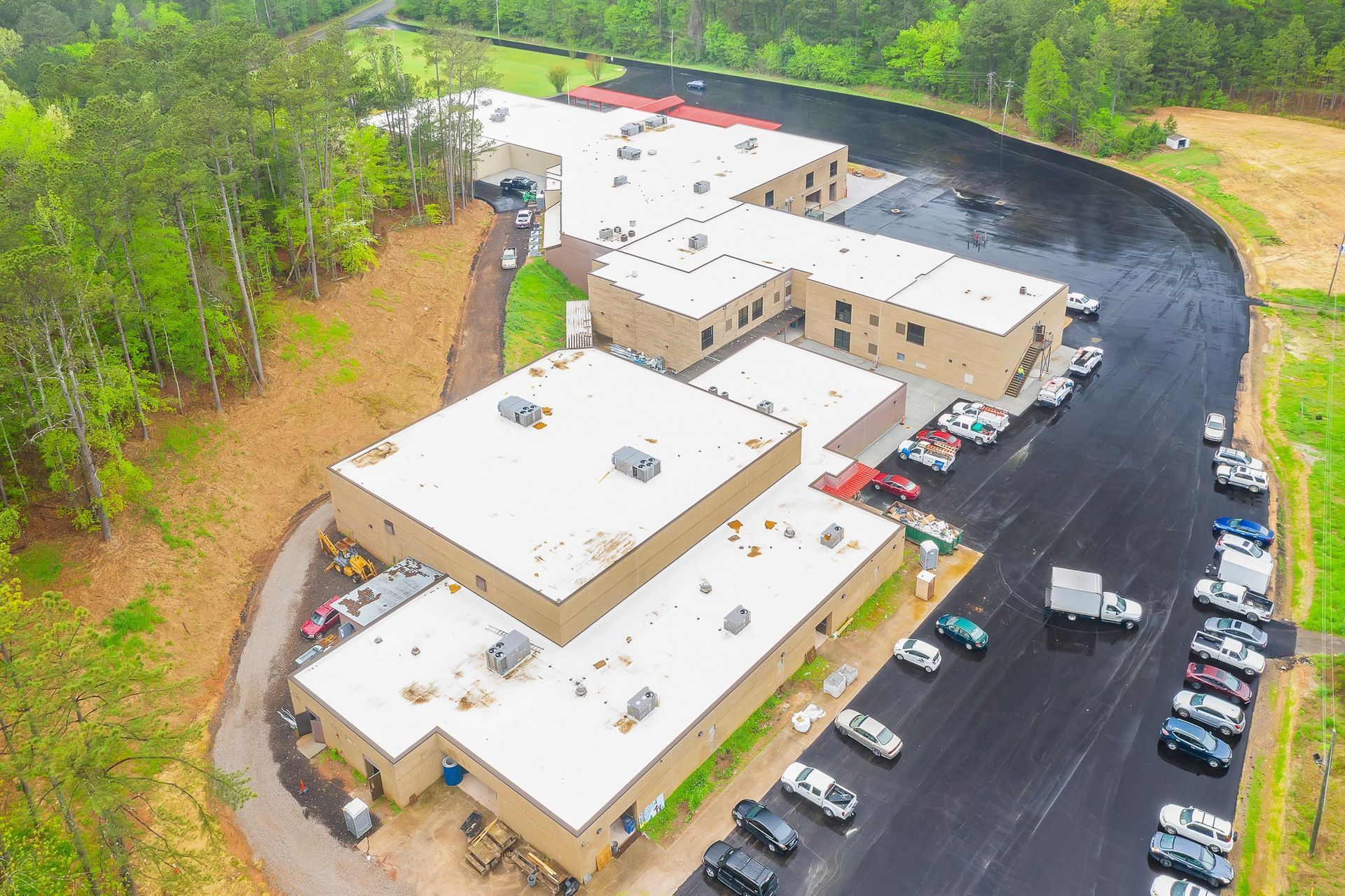Aerial view of a white industrial building complex beside a curved road and wooded area.
